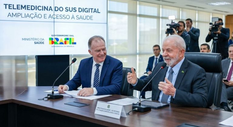 Fotografia institucional em plano medio do presidente Lula e do governador Renato Casagrande sentados lado a lado em uma mesa de reuniao de madeira escura no Ministerio da Saude em Brasilia. Ao fundo, uma grande tela de televisao exibe a escrita Telemedicina do SUS Digital Ampliacao e Acesso a Saude ao lado do logotipo oficial do Governo Federal. Lula está a direita, sorrindo e gesticulando com a mao esquerda proxima ao microfone. Casagrande esta ao centro, de terno azul e gravata listrada, olhando para Lula e sorrindo com papeis sobre a mesa. Repórteres com câmeras aparecem ao fundo.